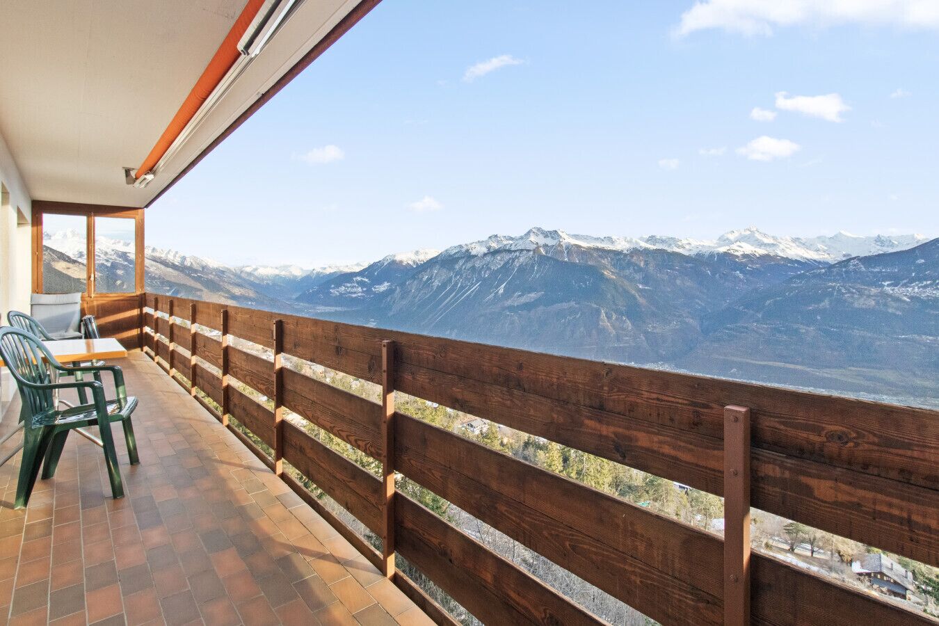Ein Balkon mit Holzwertkasten, einem Tisch und zwei Stühlen mit Blick auf eine gebirgige Landschaft mit schneebedeckten Gipfeln.