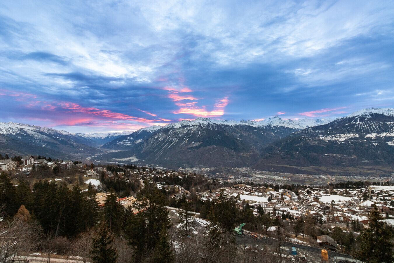 Paisaje montañoso con un pueblo en primer plano y picos nevados al fondo.