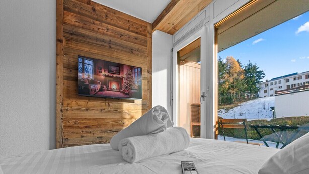 A hotel room with a wooden wall, a television, a bed with white linens, and a remote control. - Studio type chalet plein centre (Crans-Montana)