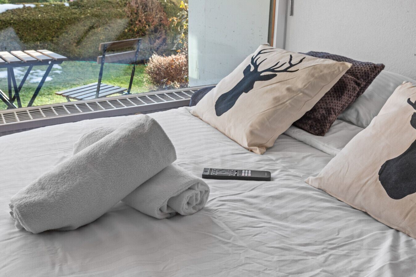 Una cama bien hecha con un control remoto, dos almohadas con motivos de ciervo y vista a un patio con mobiliario exterior.