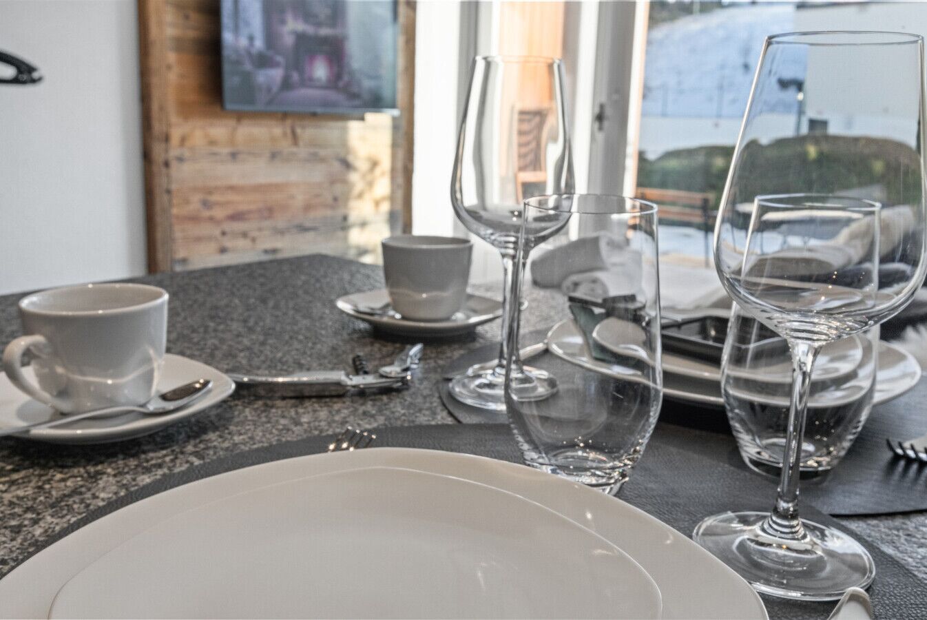 Una mesa de comedor dispuesta con platos blancos, tazas y copas de vino, con una ventana al fondo.