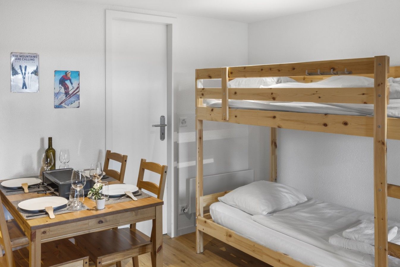Ein Schlafraum mit Etagenbetten, einem Holztisch und Stühlen, einer Tür und zwei gerahmten Bildern an der Wand.