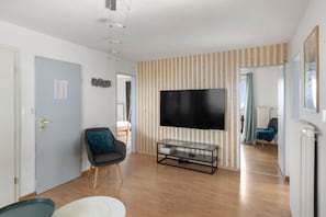 A room with a flat-screen TV mounted on a wooden wall, a chair with a cushion, a small table with a glass top, and a door with a light switch.