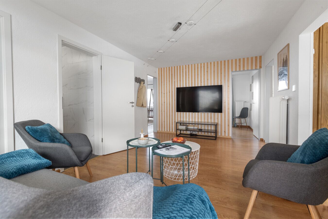Een moderne woonkamer met een flatscreen televisie, twee grijze fauteuils, een klein bijzettafeltje en een gestreept behang.