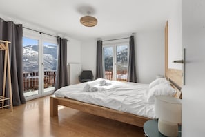 A bedroom with a wooden bed, a chair, a bedside table, and a view of mountains through large windows.