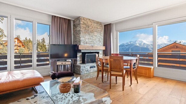 Ein modernes Wohnzimmer mit Kamin aus Stein, einem Essbereich und großen Fenstern mit Blick auf die Berge.