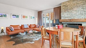 Ein modernes Wohnzimmer mit einer Ledersofa, einem Glastisch und einem Holzespeisesaal mit Stühlen. Ein Kamin aus Stein und ein großes Fenster mit Vorhängen sind ebenfalls vorhanden.