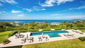 A pool area with lounge chairs and umbrellas, overlooking a coastal landscape with buildings and greenery. - La Vista #B06, gorgeous Ocean View Condo (Playa Tamarindo, Guanacaste Province)