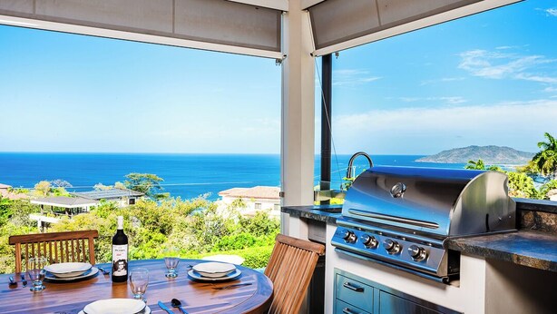 An outdoor dining area with a view of the ocean, a grill, and a table set for two. - La Vista #B06, gorgeous Ocean View Condo (Playa Tamarindo, Guanacaste Province)