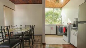 A dining area with a glass table and chairs, a kitchen with appliances, and a window with a view of greenery.