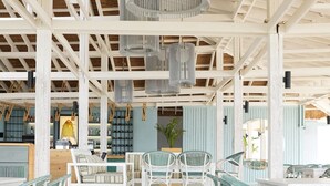 A spacious indoor area with white wooden beams and a high ceiling. There are several hanging light fixtures and a seating area with white chairs and tables. - Laguava Resort (Rumayla)