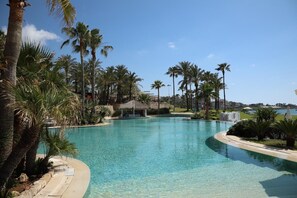 A clear blue swimming pool surrounded by palm trees and a thatched-roof structure.