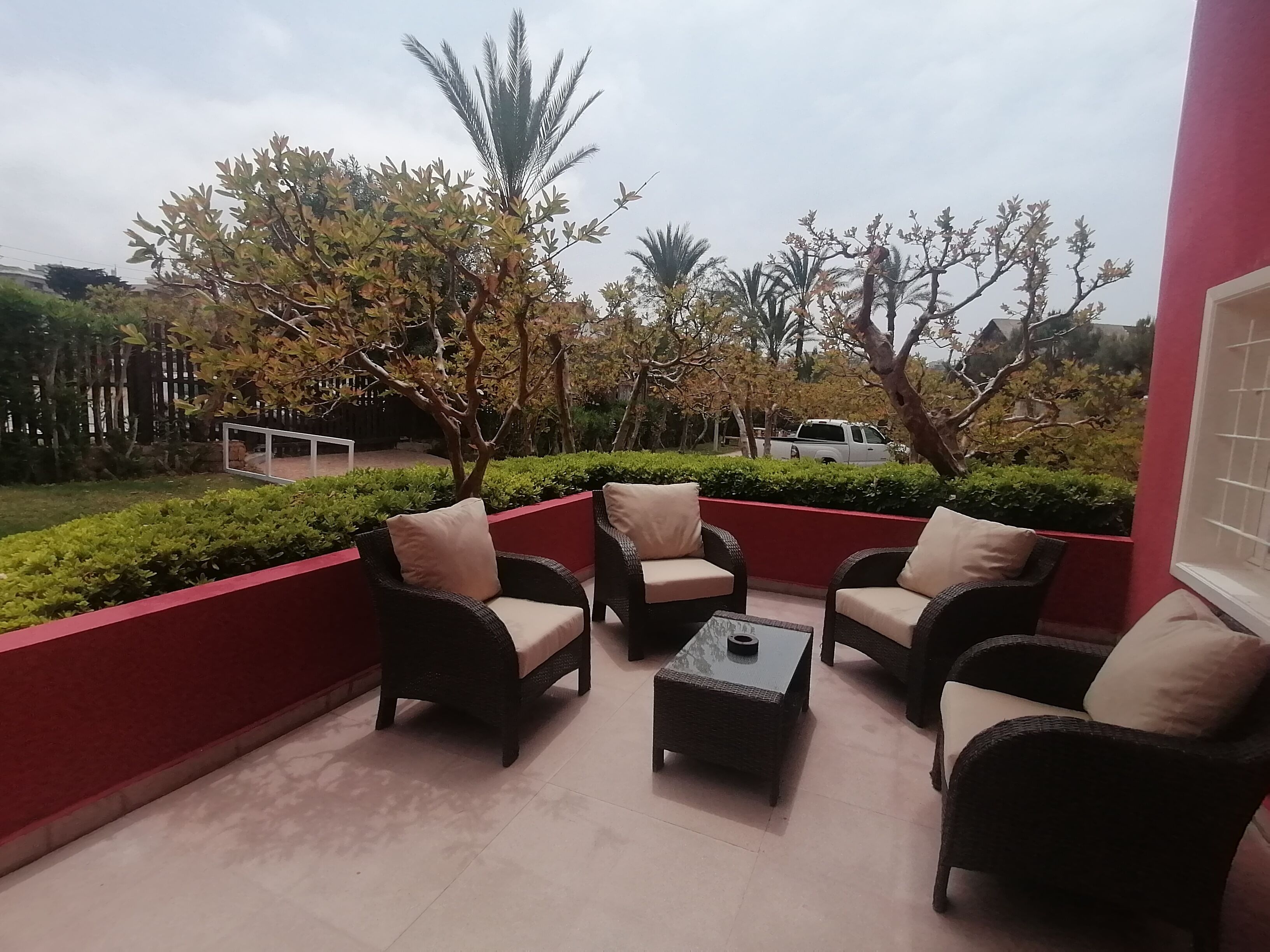 An outdoor seating area with wicker furniture and a small table, surrounded by greenery and trees.