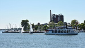 A river with a large white boat docked at the pier, surrounded by sailboats and a building with a smokestack in the background. - Apartment 24 - 1-room apartment, TV, shower (Kappeln)