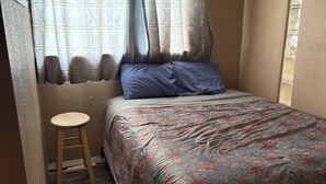 A small, cozy bedroom with a single bed, a wooden stool, and a window with patterned curtains. - Bridgeton suites (El Paso)