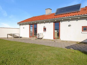 A single-story building with a red-tiled roof, a solar panel, and a patio area with a table and a fan.