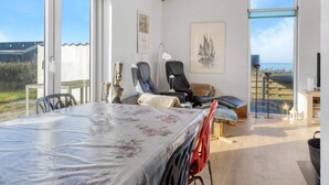 Quarto espaçoso com uma mesa grande, duas cadeiras, um sofá e uma televisão. Duas janelas oferecem vista para o mar.