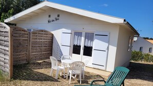 A white building with a sign reading "Nid des Pins". It has a covered patio with white plastic chairs and a green chair.
