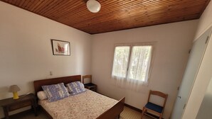 A bedroom with a wooden ceiling, a bed with floral bedding, a nightstand, a chair, and a window with curtains.