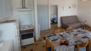 Eine kompakte Küche mit Herd, Backofen und einem weißen Kühlschrank. Ein Esstisch mit gemustertem Tischläufer und Stühlen lädt zum Verweilen ein. Im Wohnbereich befindet sich eine Couch und ein kleiner Tisch mit einer Lampe.