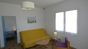 Een kamer met een gele bank, een kleine tafel met een lamp, een paarse stoel, een bed met een blauwe deken en een raam met witte gordijnen.