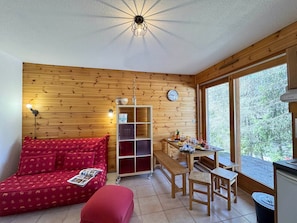 A cozy room with a wooden wall, a red sofa, a wooden table with chairs, a small bench, and a window with a view of trees. - Cozy Apartment in Vars - Pets Allowed (Vars)