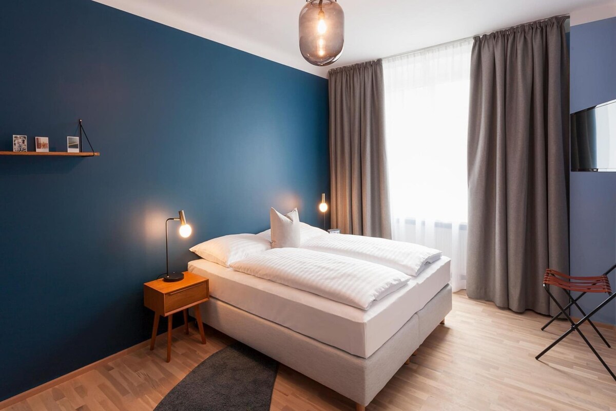 A modern bedroom with a large bed, wooden flooring, a dark blue accent wall, and a window with curtains.