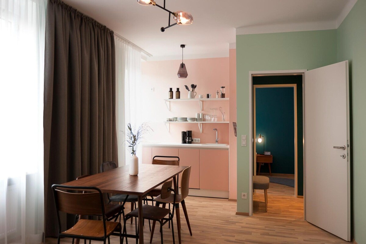 A dining area with a wooden table and chairs, a kitchen in the background, and a door leading to another room.