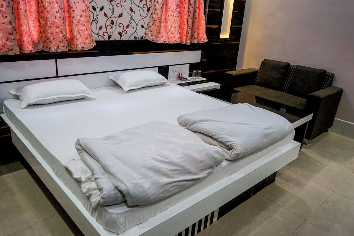 Ein ordentlich bezogenes Bett mit Kissen und Decke, ein Nachttischchen mit Lampe sowie ein Sofa in einem modernen Zimmer mit gemusterten Vorhängen.