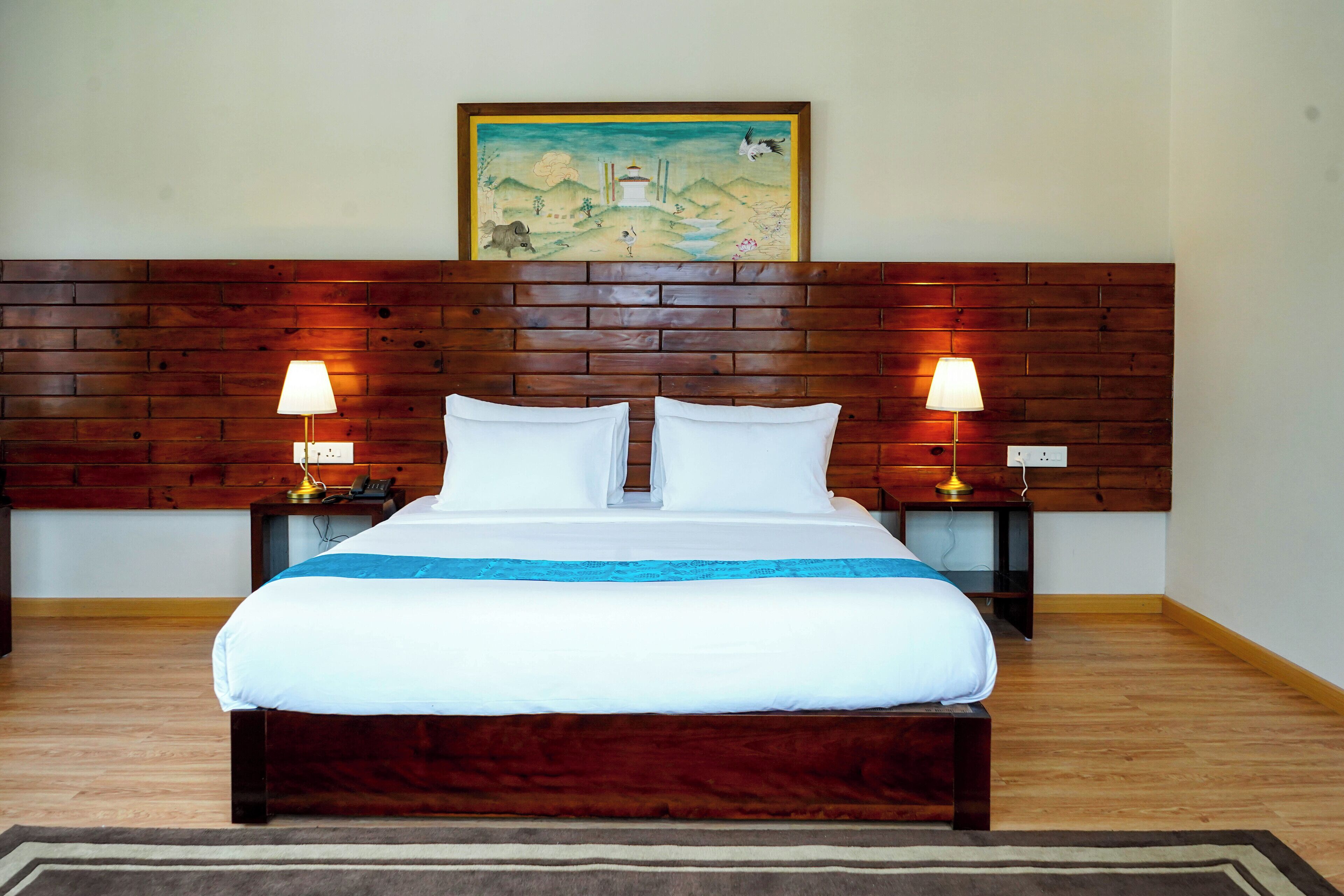 A hotel room with a wooden headboard, a large bed with white bedding, two bedside tables with lamps, and a painting above the headboard.