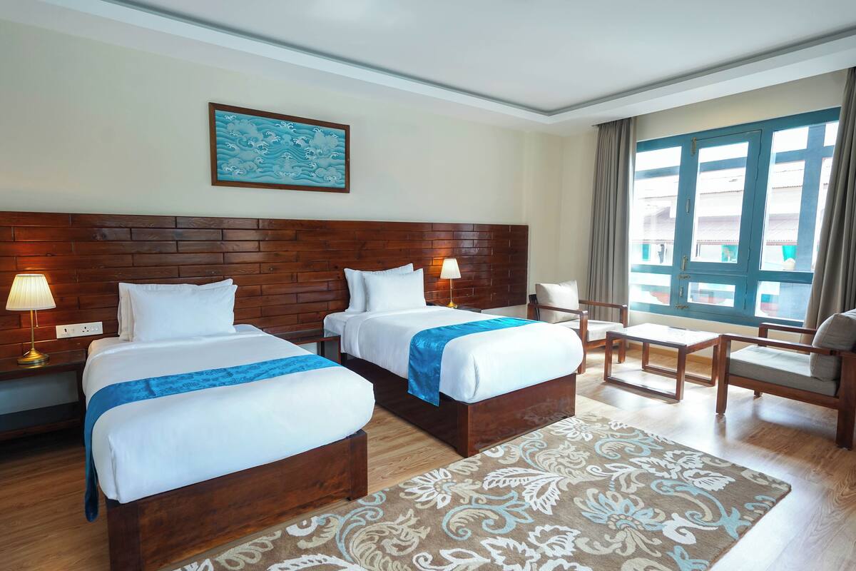 Une chambre d’hôtel avec deux lits, une tête de lit en bois, une grande fenêtre, un tapis à motifs et un tableau au mur.