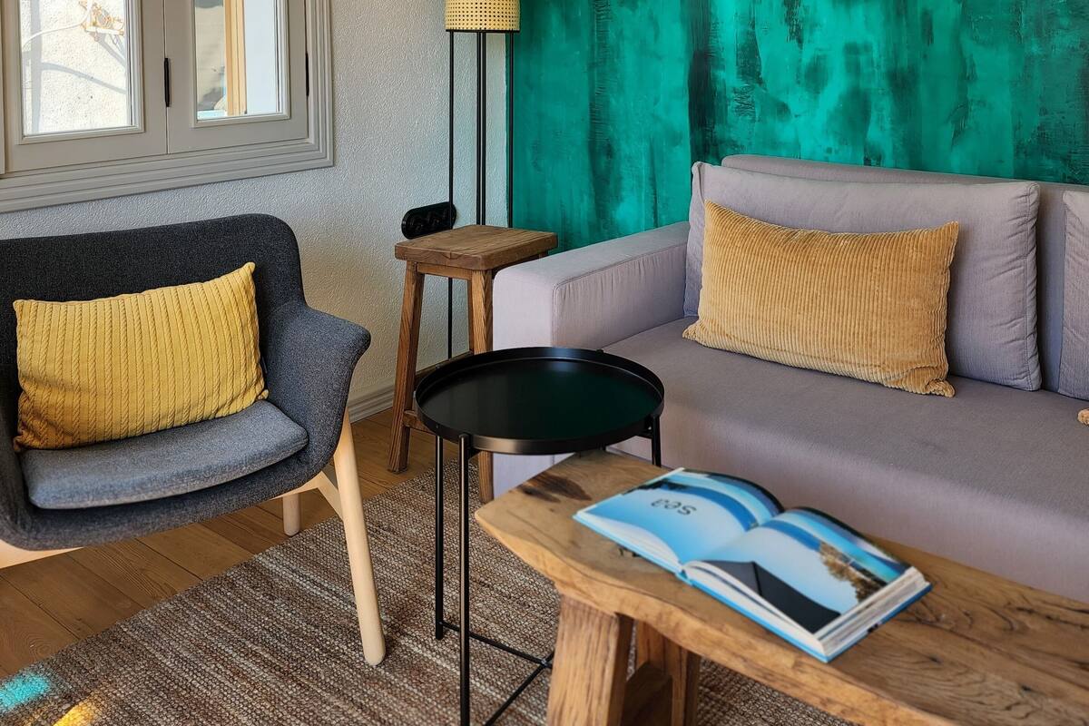 A living room with a teal accent wall, a grey sofa, a wooden coffee table, and a chair with a yellow cushion.