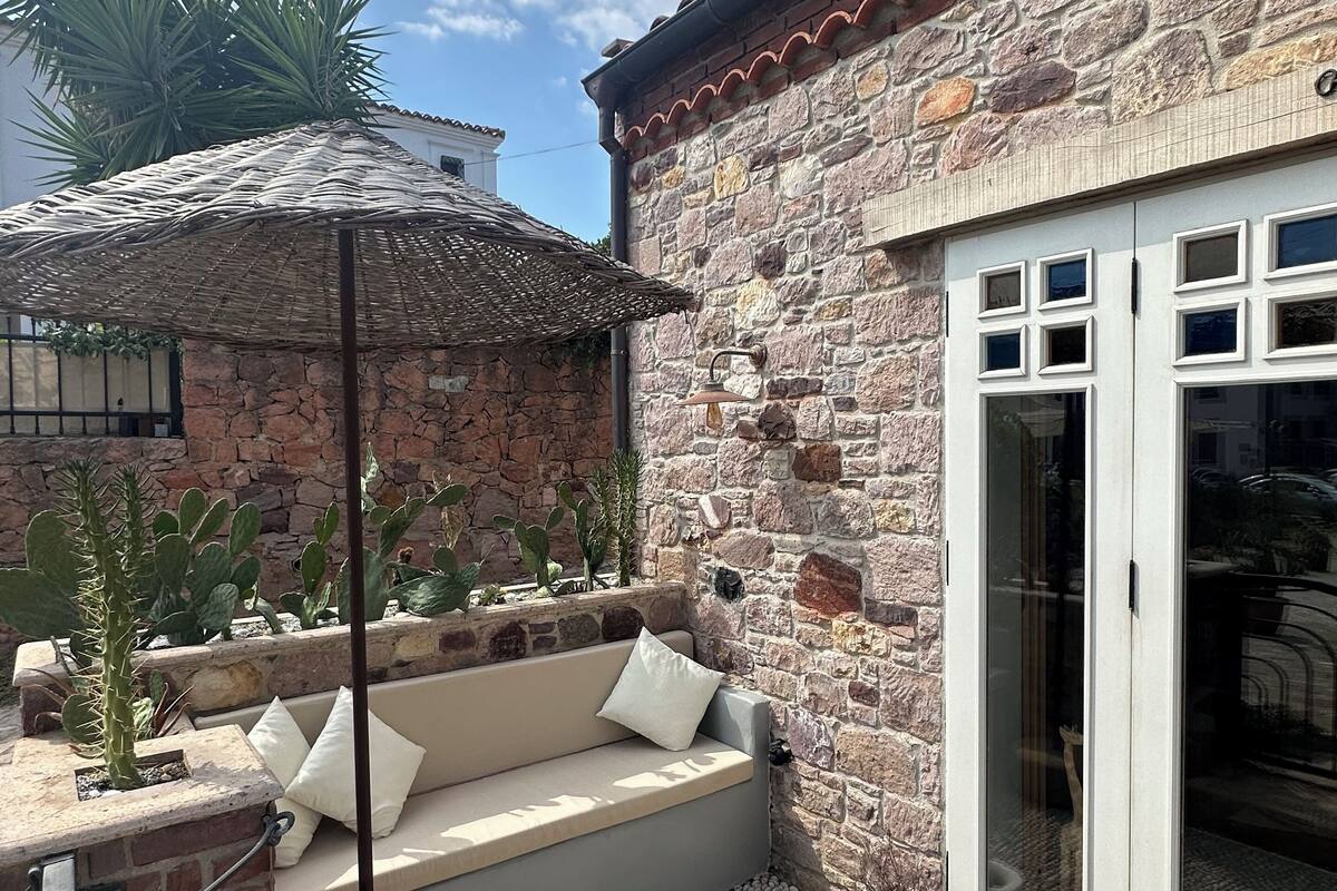 A stone building with a patio area featuring a cushioned seating arrangement, a pergola, and a variety of plants.