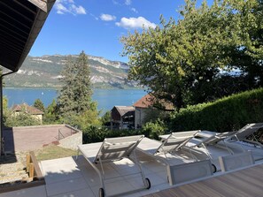 A terrace with lounge chairs overlooking a lake, a mountain backdrop, and a building with a red roof. - Villa des Lardars · Villa 4* · Panoramic lake view & close to the beach (Conjux)