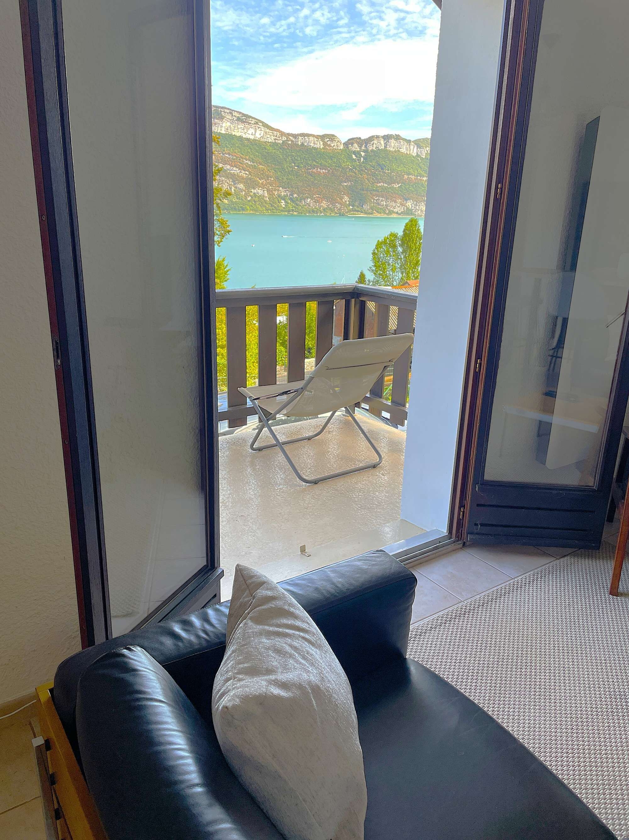 Quarto com vista para um lago e montanhas, sofá de couro preto com almofada e porta de vidro que dá para uma varanda externa com uma cadeira.