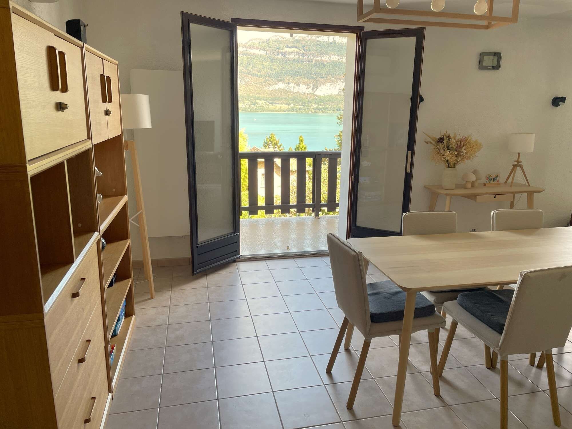 Une pièce comprenant un meuble en bois, une table à manger avec des chaises, et une porte-fenêtre coulissante donnant sur un balcon avec vue sur la mer.