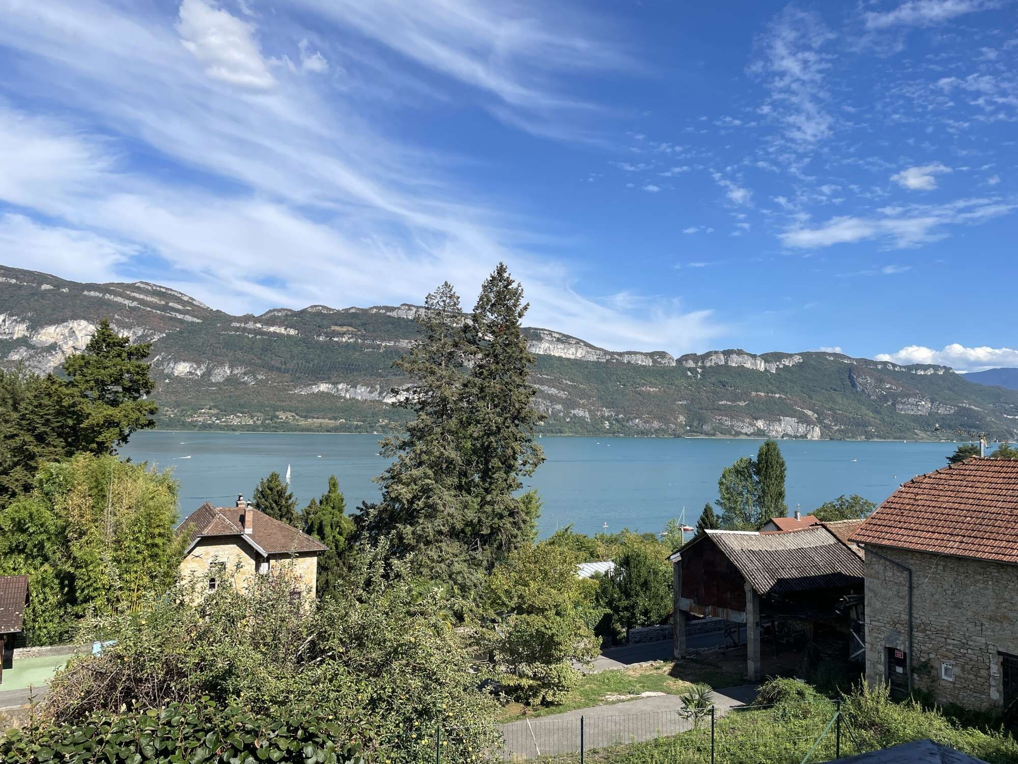 Blick auf ein Seeufer mit Gebäuden, Bäumen und Bergen im Hintergrund.
