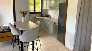 A modern kitchen with a dining area, featuring a white table, two chairs, a vase with dried flowers, and a window with a view of greenery.