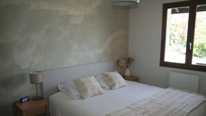 A bedroom with a bed, a nightstand with a lamp, a wall clock, and a window with a view of greenery. - Villa with panoramic lake view close to the beach (Conjux)