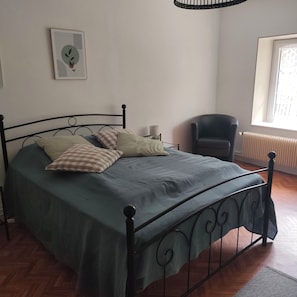 Una cama con estructura metálica, una silla, un radiador y una ventana con persianas.