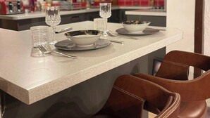 Una cocina moderna con encimera blanca, dos copas de vino, un cuenco y un plato. Hay dos sillas de cuero marrón y una lámpara colgante.