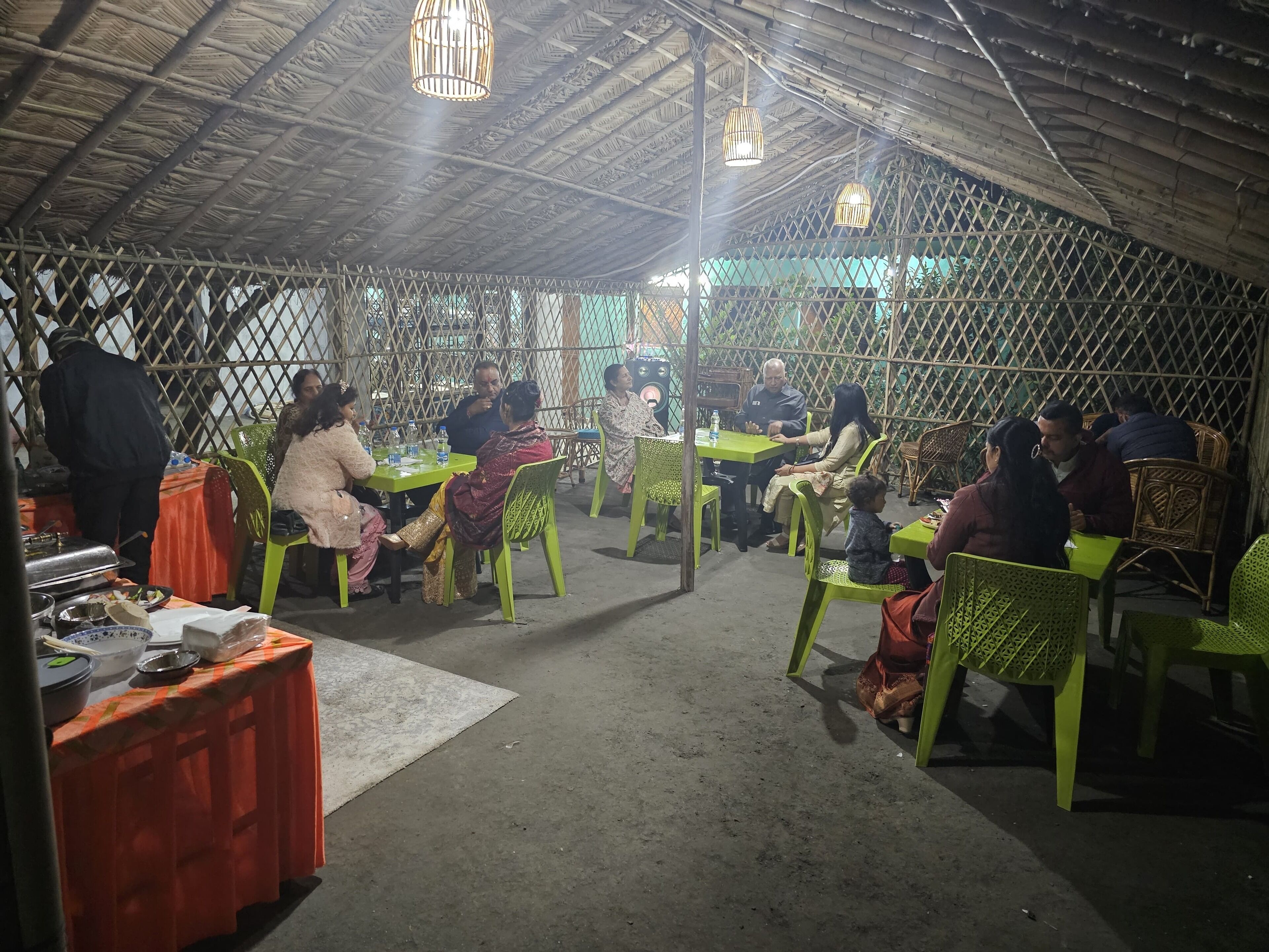 An indoor dining area with tables and chairs, a buffet table with food items, and a thatched roof structure.