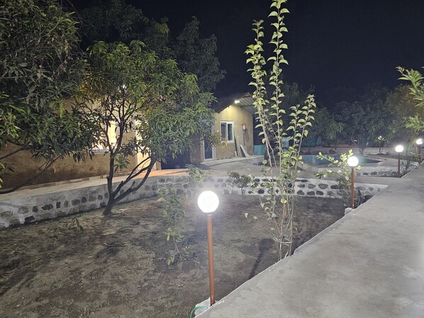 Un espace extérieur aménagé pour la nuit, avec un chemin en béton, de petits arbres et un bâtiment dont l’entrée est illuminée.