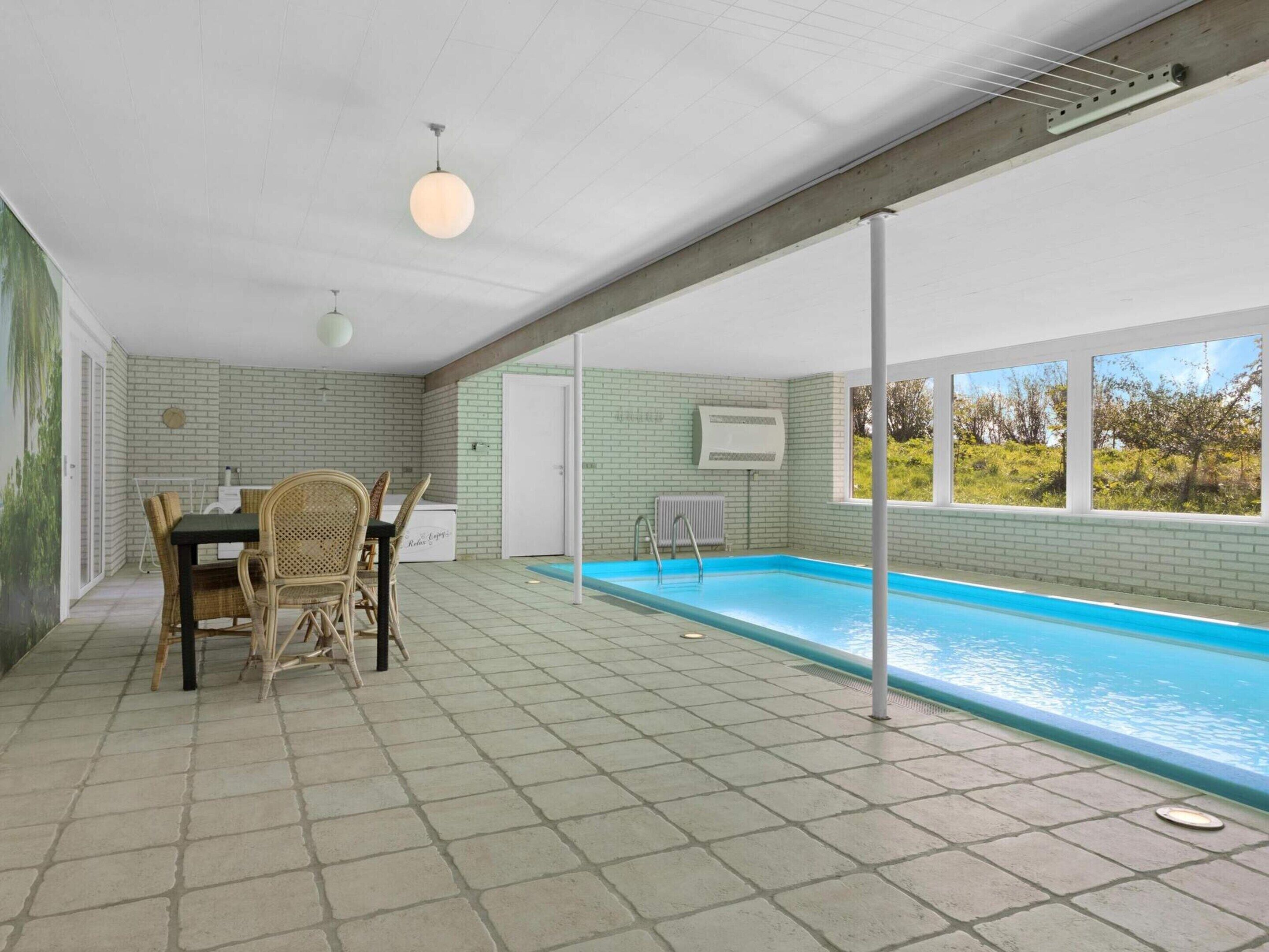Un espace autour de la piscine comprenant une table et des chaises, un radiateur et une vue sur la verdure à travers les fenêtres.
