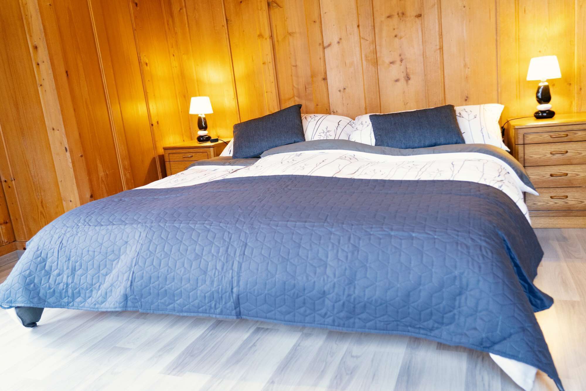 Ein Schlafzimmer mit einer Holzverkleidung, einem Bett mit blauer Bettdecke, zwei Nachttischen mit Lampen und einem zusätzlichen Nachttisch mit einer Lampe.