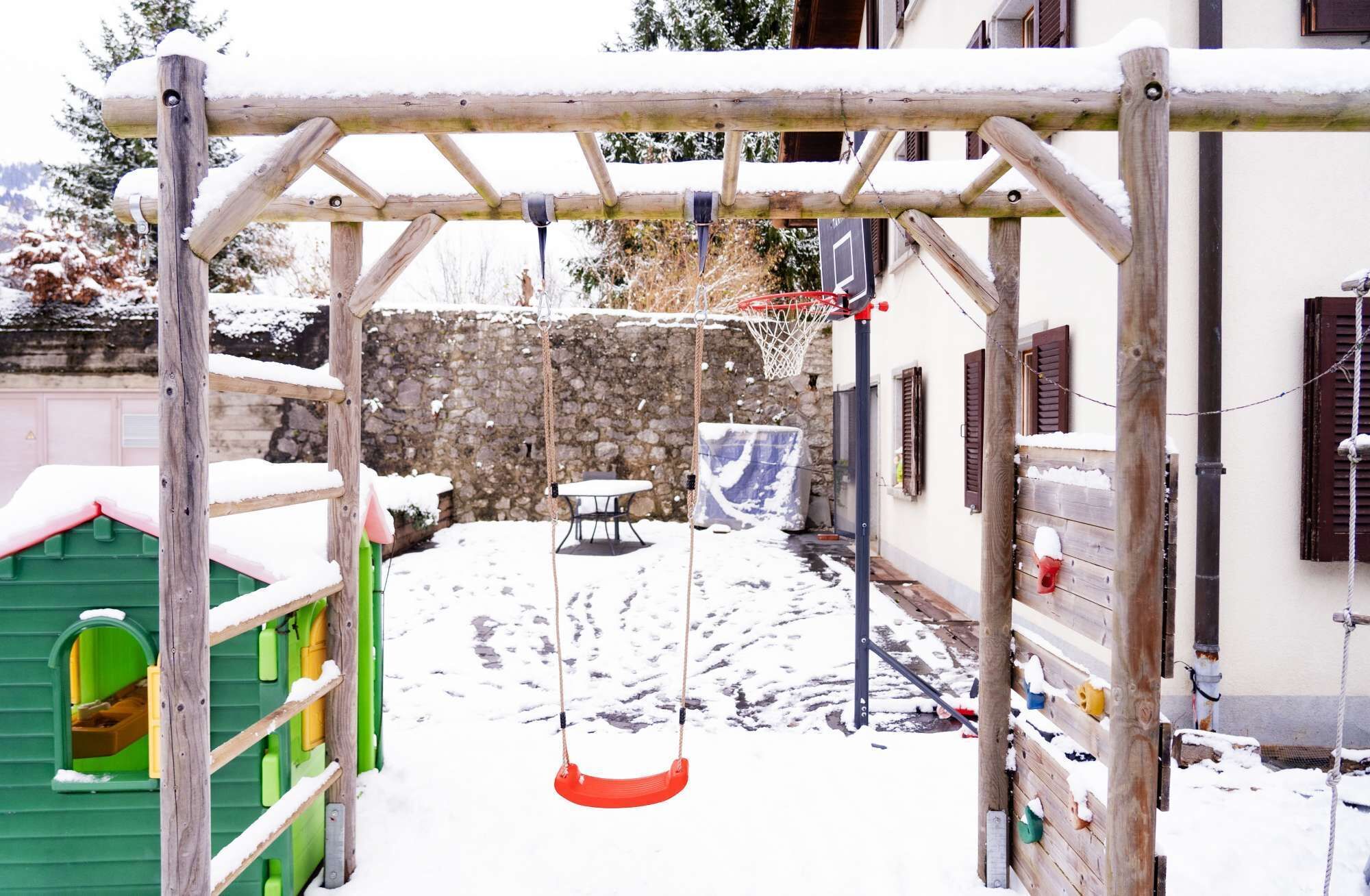 A wooden swing set in a snowy outdoor area with a stone wall and a green play structure in the background.