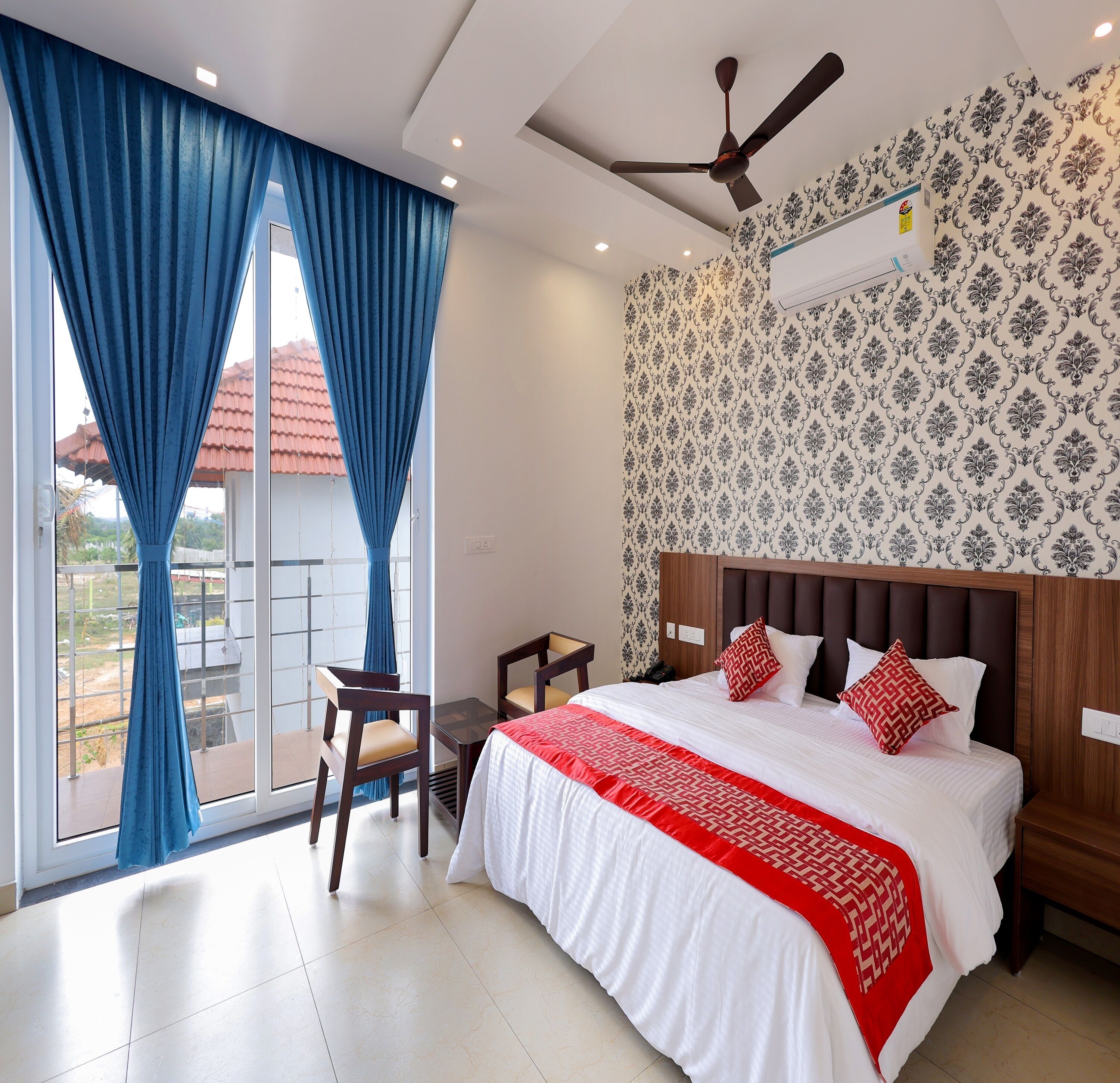 A hotel room with a bed, a desk, and a chair. There is a window with curtains, a ceiling fan, and a patterned wall.