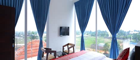 A hotel room with a bed, a desk, and a chair. There is a TV on the wall and a ceiling fan. The room has large windows with blue curtains.