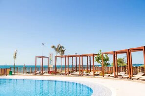 Zona junto a la piscina con sillones de descanso y pérgolas de madera, con vistas a un mar azul y cristalino.