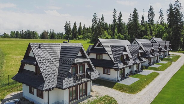 Una hilera de cabañas al estilo chalet con techos inclinados, dispuestas en línea a lo largo de un camino de grava, rodeadas por un campo cubierto de césped y un bosque frondoso.